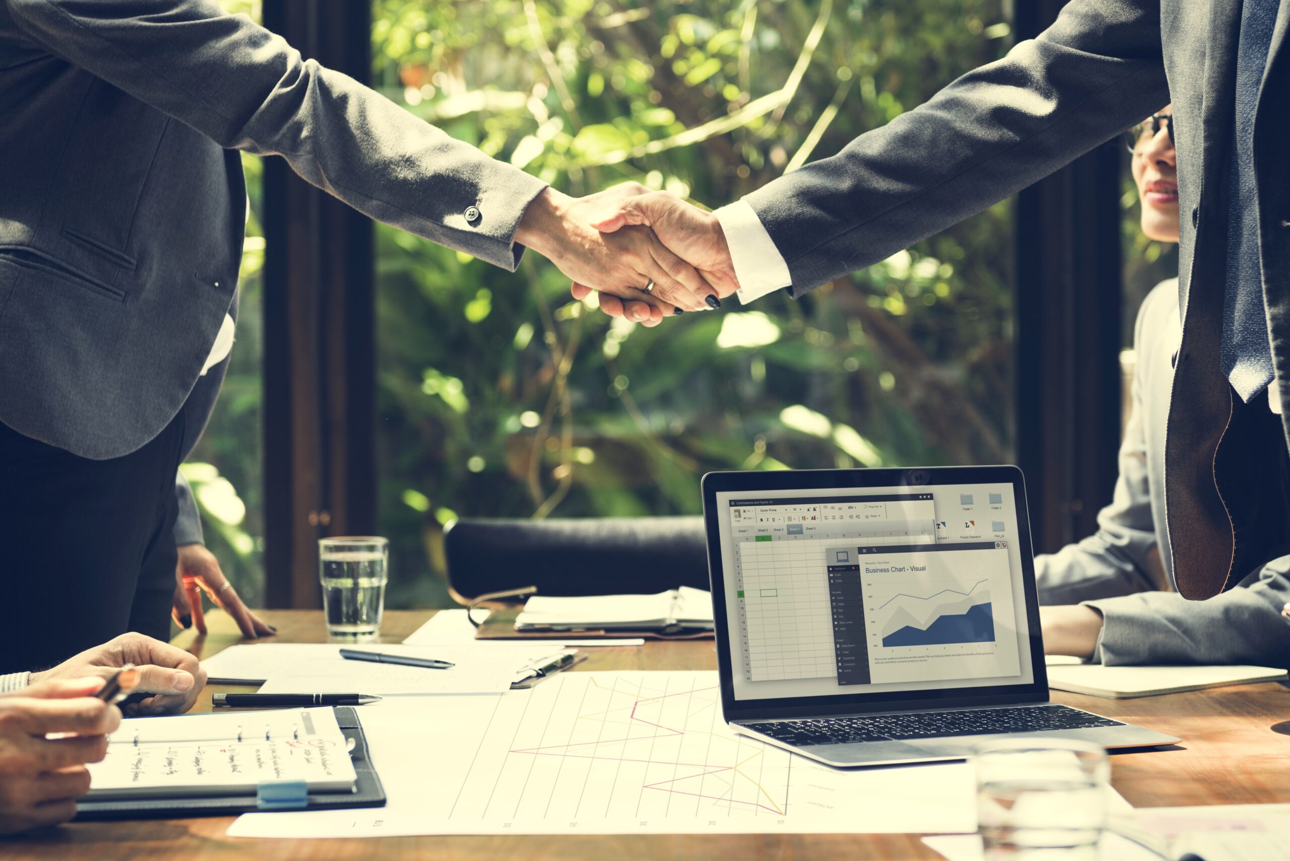 Business professionals shaking hands across a table with laptop and financial charts during a corporate meeting